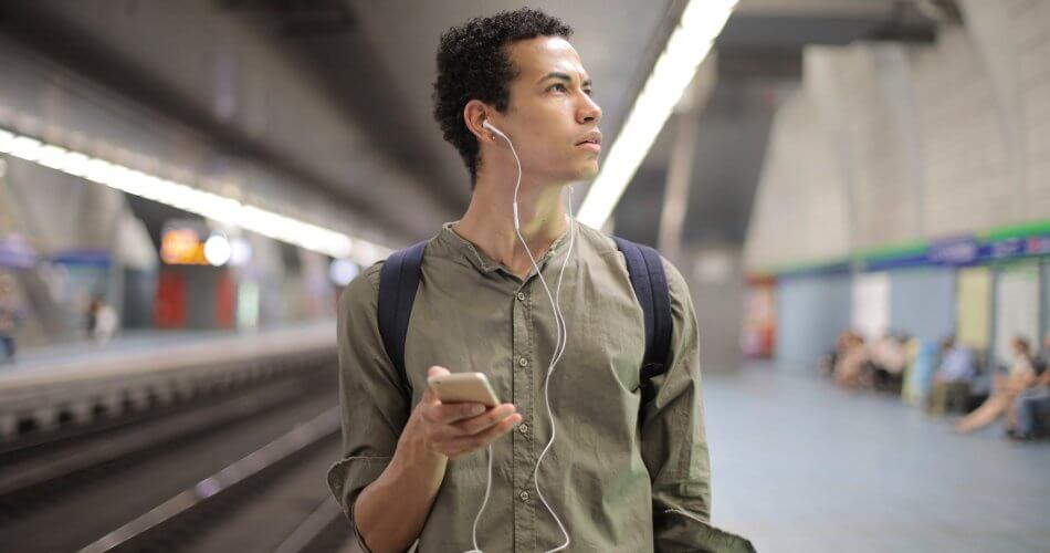 young ethnic man in earbuds listening to music while waiting for transport at contemporary subway station
