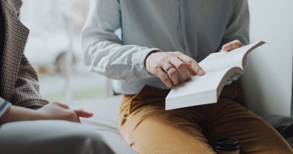 man in white dress shirt and brown pants sitting on white chair reading book